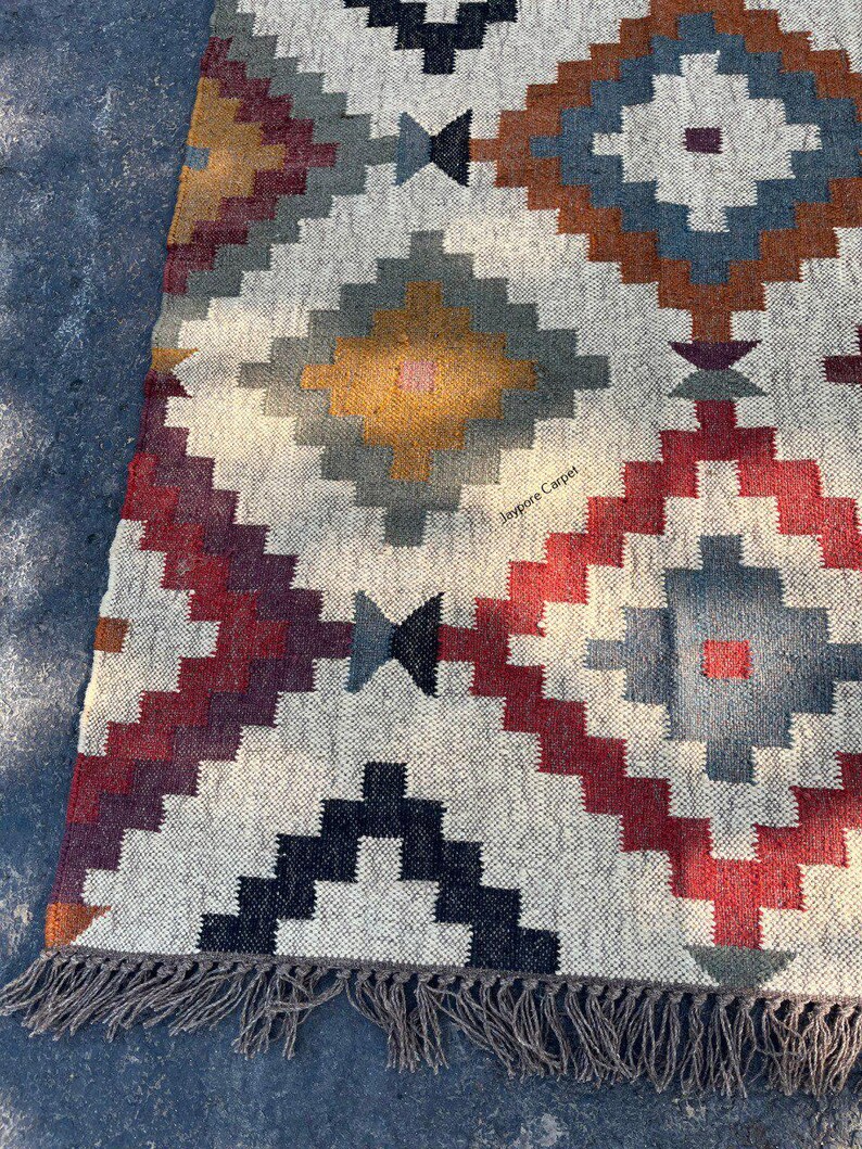May include: A rectangular, woven rug with a geometric pattern in shades of red, blue, brown, and yellow on a cream background. The rug has a fringe along one edge and the text "Jaypore Carpet" is visible.