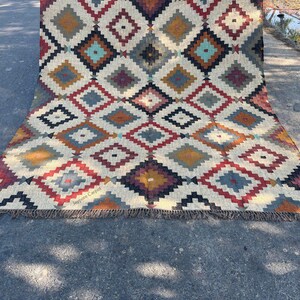 May include: A large, rectangular rug with a geometric diamond pattern in shades of red, blue, yellow, and brown on a cream background. The rug has a fringed edge and the text "Jaypore Carpet" is visible.