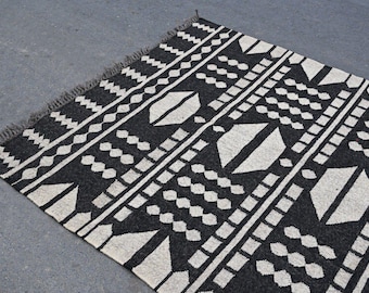 May include: A black and white patterned rug with a geometric design. The rug features repeating shapes, including diamonds, rectangles, and lines. The rug has a fringed edge and is made of a woven material.