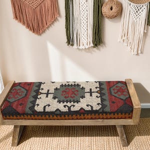 May include: A wooden bench with a rectangular seat featuring a patterned rug design in red, black, gray, and cream colors. The bench has a natural wood frame and is placed on a woven jute rug. Decorative wall hangings are visible in the background.