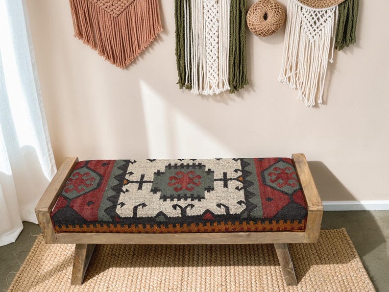 May include: A wooden bench with a rectangular seat featuring a patterned rug design in red, black, gray, and cream colors. The bench has a natural wood frame and is placed on a woven jute rug. Decorative wall hangings are visible in the background.