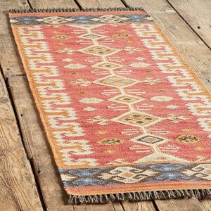 May include: A rectangular rug with a coral-colored background and a geometric pattern in white, brown, and yellow. The rug has a blue and brown border and fringed edges. The rug is placed on a wooden surface.