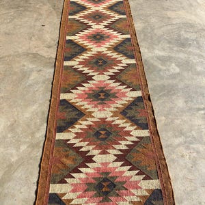 May include: A long, narrow runner rug with a geometric diamond pattern in shades of brown, pink, cream, and navy blue. The rug has a fringed edge and is likely made of natural fibers. The design is reminiscent of Southwestern or tribal styles.