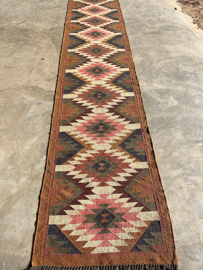 May include: A long, narrow runner rug with a geometric diamond pattern in shades of brown, pink, cream, and navy blue. The rug has a fringed edge and is likely made of natural fibers. The design is reminiscent of Southwestern or tribal styles.