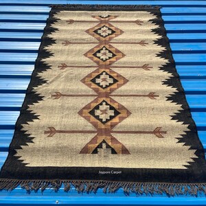 May include: Rectangular beige rug with a geometric pattern in brown, black, and gold. The rug has a black border with fringe and arrow-like designs. The text "Jaypore Carpet" is visible.