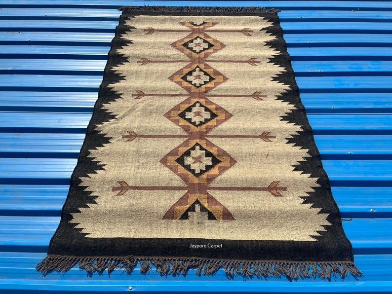 May include: Rectangular beige rug with a geometric pattern in brown, black, and gold. The rug has a black border with fringe and arrow-like designs. The text "Jaypore Carpet" is visible.