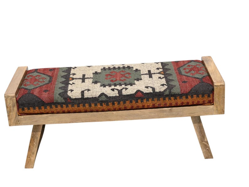 May include: A wooden bench with a rectangular seat featuring a colorful, patterned rug design in red, green, black, and cream. The bench has light-colored wooden legs and armrests, creating a rustic aesthetic.