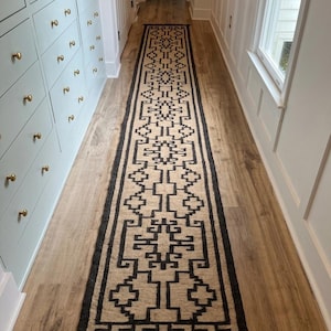 Puede incluir: Una alfombra larga y rectangular con un patrón geométrico en negro y beige. La alfombra tiene flecos en los extremos y el texto "Jaypore Carpet" es visible. La alfombra está en un pasillo con suelo de madera.