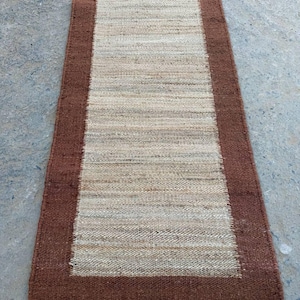 May include: A rectangular runner rug featuring a woven texture with a wide, solid dark brown border framing a lighter, natural beige center panel. The rug has a consistent, coarse fiber construction throughout its entire surface area.