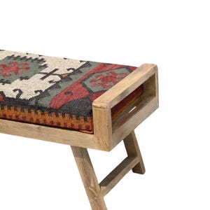 May include: Wooden bench with a rectangular seat featuring a patterned textile in shades of beige, red, green, and black. The bench has a natural wood finish and angled legs, suitable for indoor use.