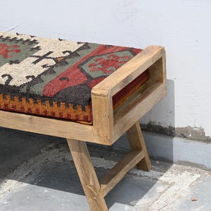 May include: Wooden bench with a rectangular seat featuring a patterned textile in shades of red, green, and beige. The bench has a natural wood finish and a simple, geometric design. The legs are angled for stability.