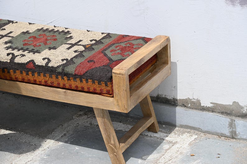 May include: Wooden bench with a rectangular seat featuring a patterned textile in shades of red, green, and beige. The bench has a natural wood finish and a simple, geometric design. The legs are angled for stability.