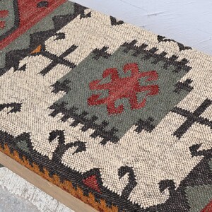 May include: A rectangular bench with a light wood frame and a patterned textile top. The textile features a central design in cream, gray, and red, with black and orange accents. The overall design has a geometric, tribal aesthetic.