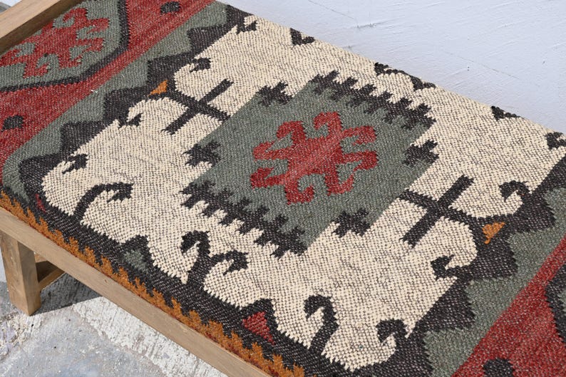 May include: A rectangular bench with a light wood frame and a patterned textile top. The textile features a central design in cream, gray, and red, with black and orange accents. The overall design has a geometric, tribal aesthetic.