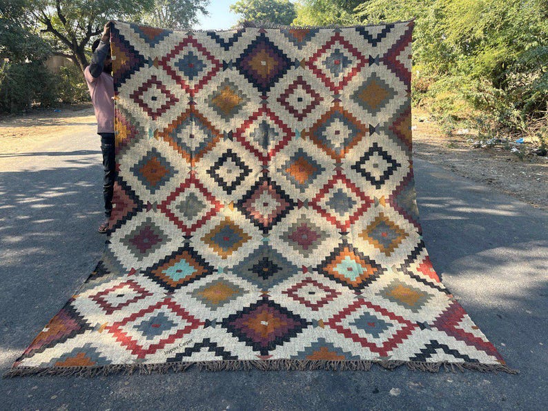May include: A large, rectangular rug with a geometric diamond pattern in shades of cream, red, blue, orange, and black. The rug has a fringed edge and is displayed outdoors. The text "Jaipur Carpet" is visible on the rug.