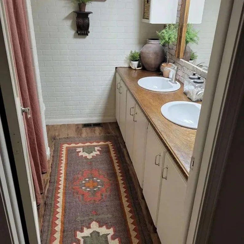 Pode incluir: Uma casa de banho com um tapete comprido e estampado em tons de cinza, vermelho e laranja. O quarto apresenta uma parede de tijolo branco, uma bancada de madeira com duas pias brancas e arm&aacute;rios brancos. Um espelho e itens decorativos s&atilde;o vis&iacute;veis.