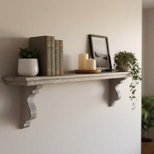May include: A wall-mounted wooden shelf featuring a weathered gray finish and two decorative corbel brackets. The shelf holds a white ceramic pot with a succulent, four hardcover books, two cylindrical pillar candles, and a framed photograph.