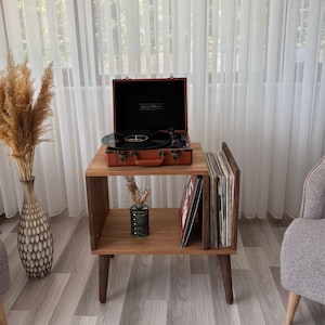 May include: A wooden record player stand with a brown suitcase-style record player on top. The stand has a lower shelf for vinyl record storage. A vase with dried plants and a decorative vase are also visible.