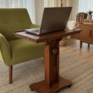 May include: A dark brown wooden side table with a rectangular top and a central vertical support column featuring a round adjustment knob. The table has a wide, stable base with small casters and supports a grey laptop and a white ceramic mug.