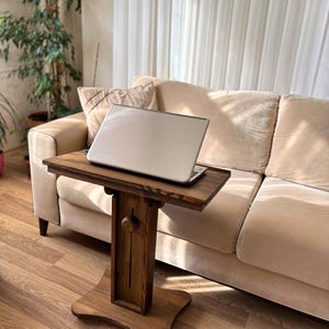 May include: A wooden laptop table with a dark brown finish. The table has a rectangular top and a single pedestal base. A silver laptop is placed on the table. The table is positioned in front of a beige sofa.