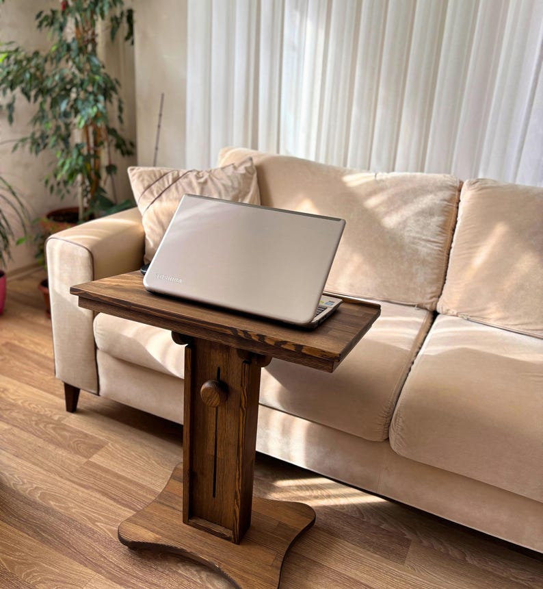 May include: A wooden laptop table with a dark brown finish. The table has a rectangular top and a single pedestal base. A silver laptop is placed on the table. The table is positioned in front of a beige sofa.