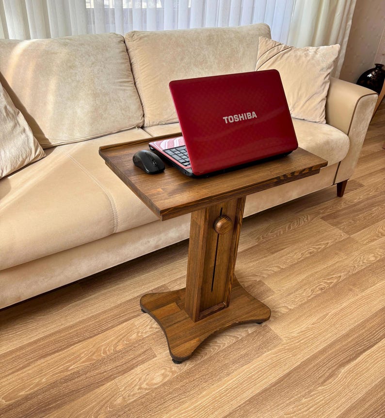 May include: A wooden laptop table with a red Toshiba laptop and a black mouse on top. The table is brown and has a decorative base. The laptop is open, displaying the keyboard. The table is next to a beige sofa.