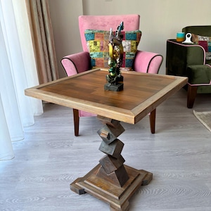 May include: A square wooden side table with a dark brown tabletop and a lighter brown frame. The base is a unique design of stacked geometric shapes. A decorative statue sits on the table, with a pink armchair in the background.