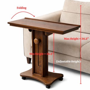 May include: A brown wooden folding side table with adjustable height. The table has a maximum height of 36.2 inches and a minimum height of 24.4 inches. It is on wheels and is positioned next to a beige sofa.