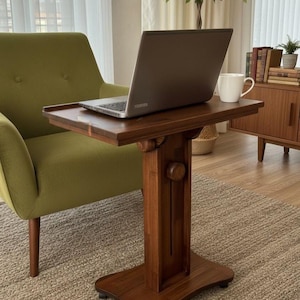 May include: A dark brown wooden side table with a rectangular top, a central vertical support column featuring a circular adjustment knob, and a wide base equipped with small casters for mobility, supporting a grey laptop and a white ceramic mug.