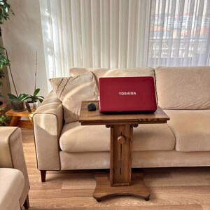May include: A red Toshiba laptop sits on a small, dark wooden table with a mouse. The table is in front of a beige sofa. The laptop is open, and the screen is facing the viewer. The table has a decorative design.