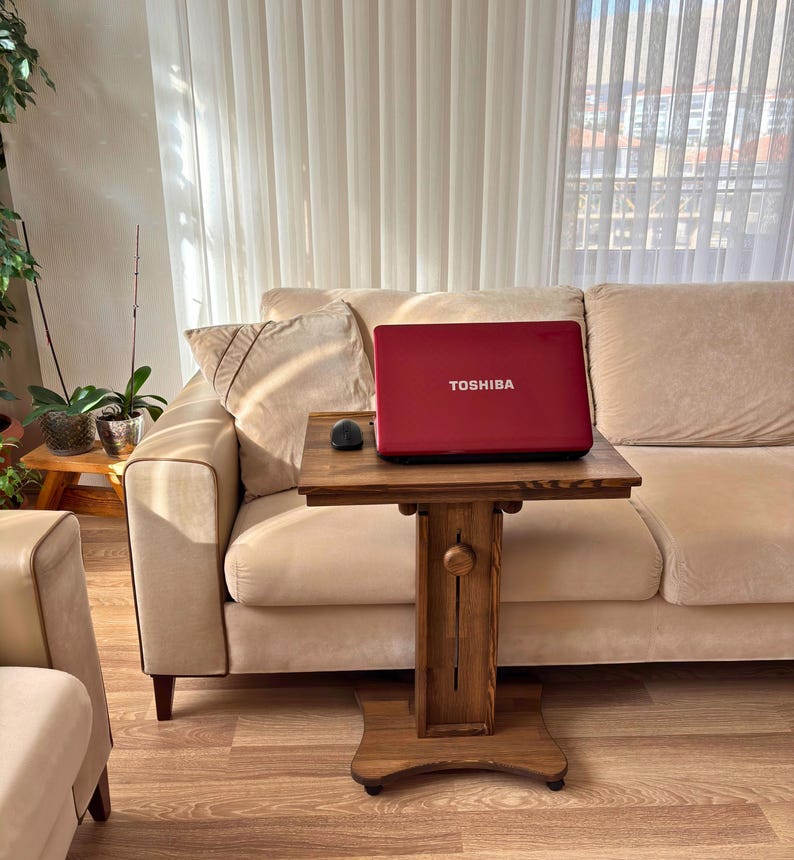 May include: A red Toshiba laptop sits on a small, dark wooden table with a mouse. The table is in front of a beige sofa. The laptop is open, and the screen is facing the viewer. The table has a decorative design.