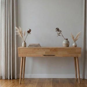 May include: A light-toned wooden console table with four tapered legs and a single drawer featuring a thin black handle. On top sit three ceramic vases containing dried pampas grass and protea flowers, alongside a small stack of books.