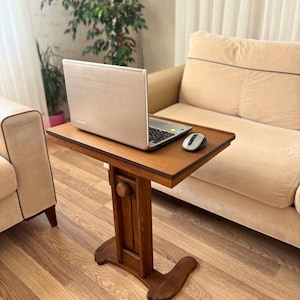 May include: A wooden laptop table with a rectangular top and a decorative base. A silver laptop and a computer mouse are on the table. The table is made of dark brown wood and is designed to be placed next to a sofa or chair.