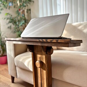 May include: A wooden side table with a laptop on top. The table is made of dark stained wood and has a decorative design. The laptop is silver and open. The table is next to a beige sofa.