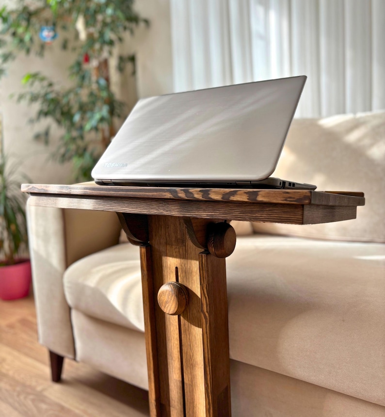 May include: A wooden side table with a laptop on top. The table is made of dark stained wood and has a decorative design. The laptop is silver and open. The table is next to a beige sofa.