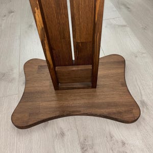 May include: A wooden stand with a dark brown finish. The stand has a wide, curved base and a vertical support with a central slot. The wood grain is visible, adding texture to the piece. The stand appears to be designed for holding an item.