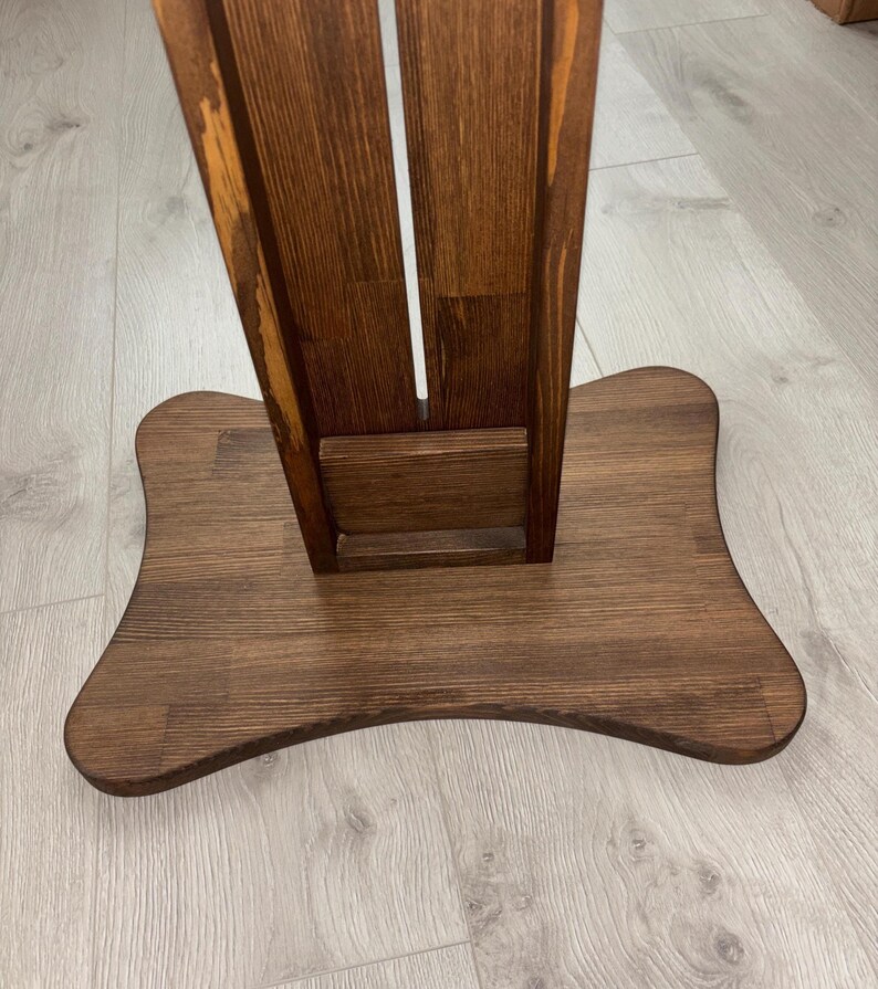 May include: A wooden stand with a dark brown finish. The stand has a wide, curved base and a vertical support with a central slot. The wood grain is visible, adding texture to the piece. The stand appears to be designed for holding an item.