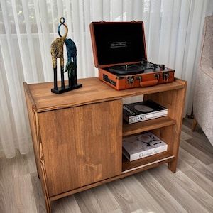 May include: A wooden cabinet with a sliding door and open shelves. On top sits a vintage orange record player and a decorative sculpture. Books with the titles "CHANEL" and "Christian Dior" are on the shelves.