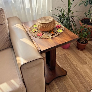 May include: A small, dark brown wooden side table with a square top and a decorative base. A woven basket with a lid sits on top of a woven placemat decorated with colorful flower shapes. The table is next to a beige sofa.