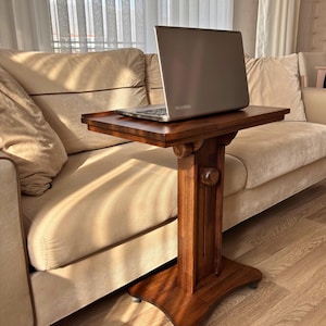 May include: A brown wooden side table with a laptop on top. The table has a square top and a decorative column base. The laptop is silver and has the brand name "Toshiba" on it. The table is placed next to a beige sofa.