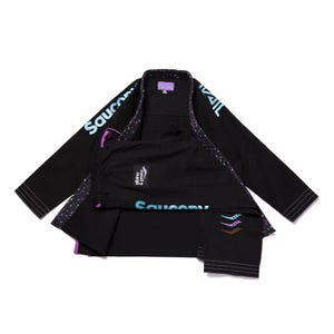 Pode incluir: Uniforme preto de Jiu-Jitsu com o logotipo Saucony em verde-azulado e roxo. O uniforme inclui um casaco e calças, com as palavras "Saucony" e "Brazil" impressas no casaco. As calças têm o logotipo "Saucony" e três listras coloridas.