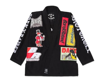 May include: Black Jiu-Jitsu gi with multiple patches. Patches include "Born x Raised" in red and white, a cartoon fox in a top hat, and "Shoyoroll" in yellow. The gi has "Born x Raised" text on the sleeves.