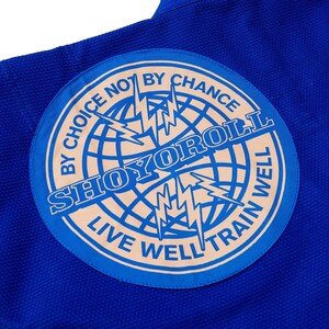 May include: Close-up of a blue martial arts uniform with a circular patch. The patch features the text "SHOYOROLL" in a stylized font, surrounded by the phrases "BY CHOICE NOT BY CHANCE" and "LIVE WELL TRAIN WELL". A globe and lightning bolts are also featured.