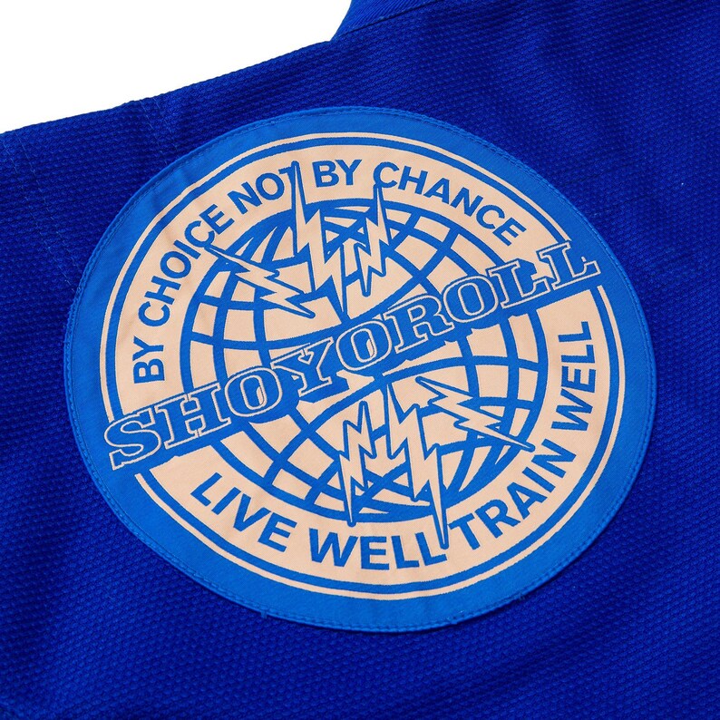 May include: Close-up of a blue martial arts uniform with a circular patch. The patch features the text "SHOYOROLL" in a stylized font, surrounded by the phrases "BY CHOICE NOT BY CHANCE" and "LIVE WELL TRAIN WELL". A globe and lightning bolts are also featured.