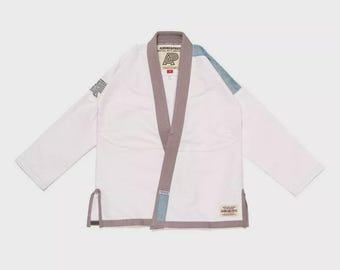 May include: White martial arts uniform with a gray collar and trim. The uniform has a blue accent on the shoulder and a logo on the chest. The brand name "ALBINO & PRETO" is printed on the collar and the bottom hem.