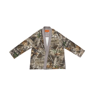 Puede incluir: Una chaqueta de manga larga estilo kimono con un patrón de camuflaje de bosque realista en tonos marrón, beige y verde. La prenda tiene un cuello grueso de color gris claro sólido y una etiqueta interior naranja en el cuello con texto.