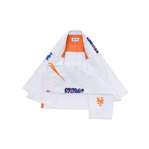 Op de afbeelding: Wit vechtsportuniform met oranje en blauwe accenten. De jas en broek tonen het woord "Mets" in blauw en het teamlogo in oranje. Het uniform is plat uitgespreid.