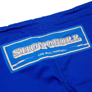 May include: Close-up of blue martial arts uniform with a rectangular patch. The patch features the brand name "SHOYOROLL" in a stylized font, with the text "LIVE WELL TRAIN WELL" below it. The uniform is made of a durable fabric.