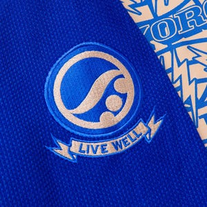 May include: Close-up of a blue fabric with a circular emblem. The emblem features a stylized design in beige and blue, with the words "LIVE WELL" on a banner below. A portion of a patterned fabric with blue text is visible.