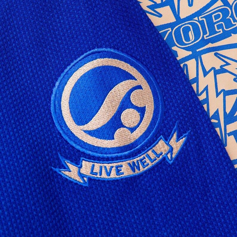 May include: Close-up of a blue fabric with a circular emblem. The emblem features a stylized design in beige and blue, with the words "LIVE WELL" on a banner below. A portion of a patterned fabric with blue text is visible.